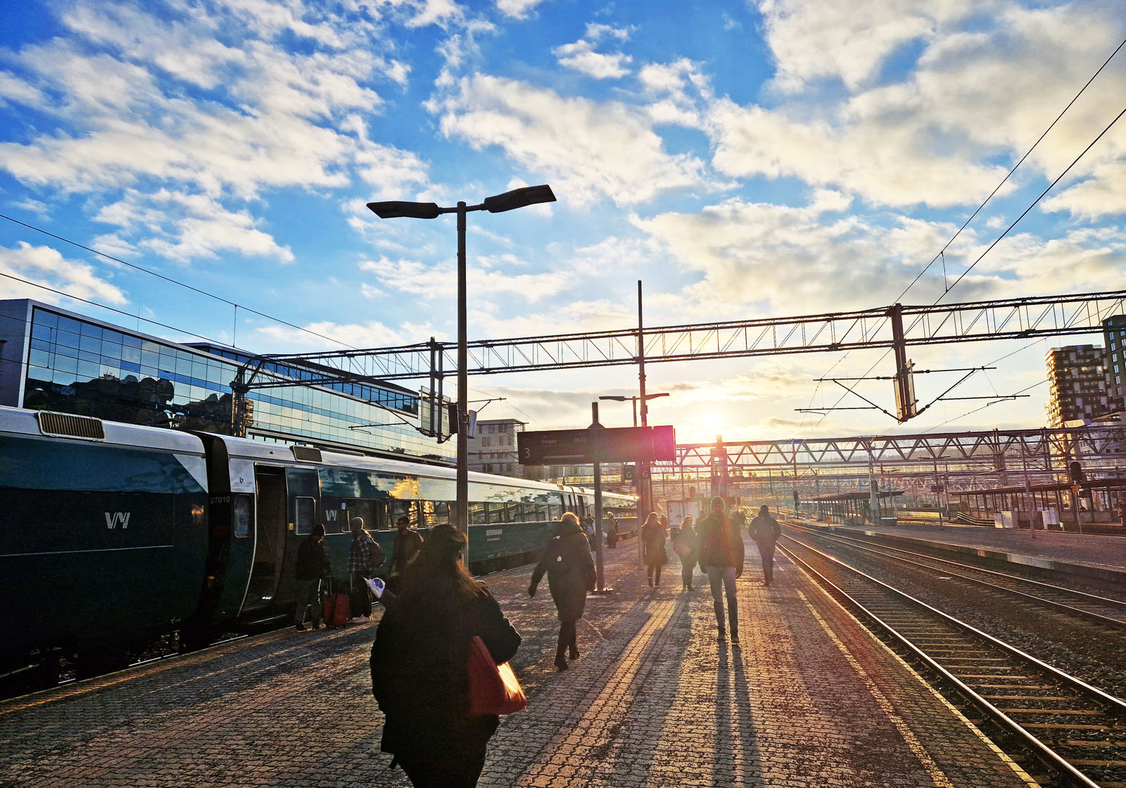 Lav sol over perrongene på Oslo S. Folk går på plattformen mot sola. Et tog står på ett av sporene.