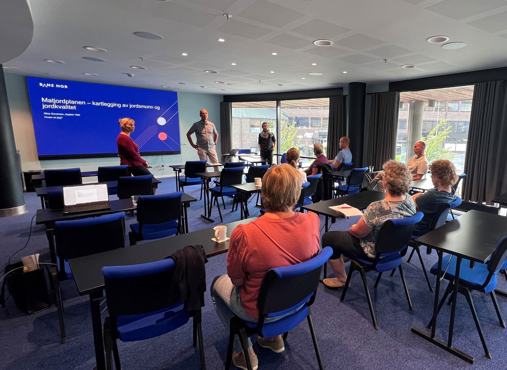 Kvinner og menn i sal. Foran en skjerm som viser presentasjon og tre personer.