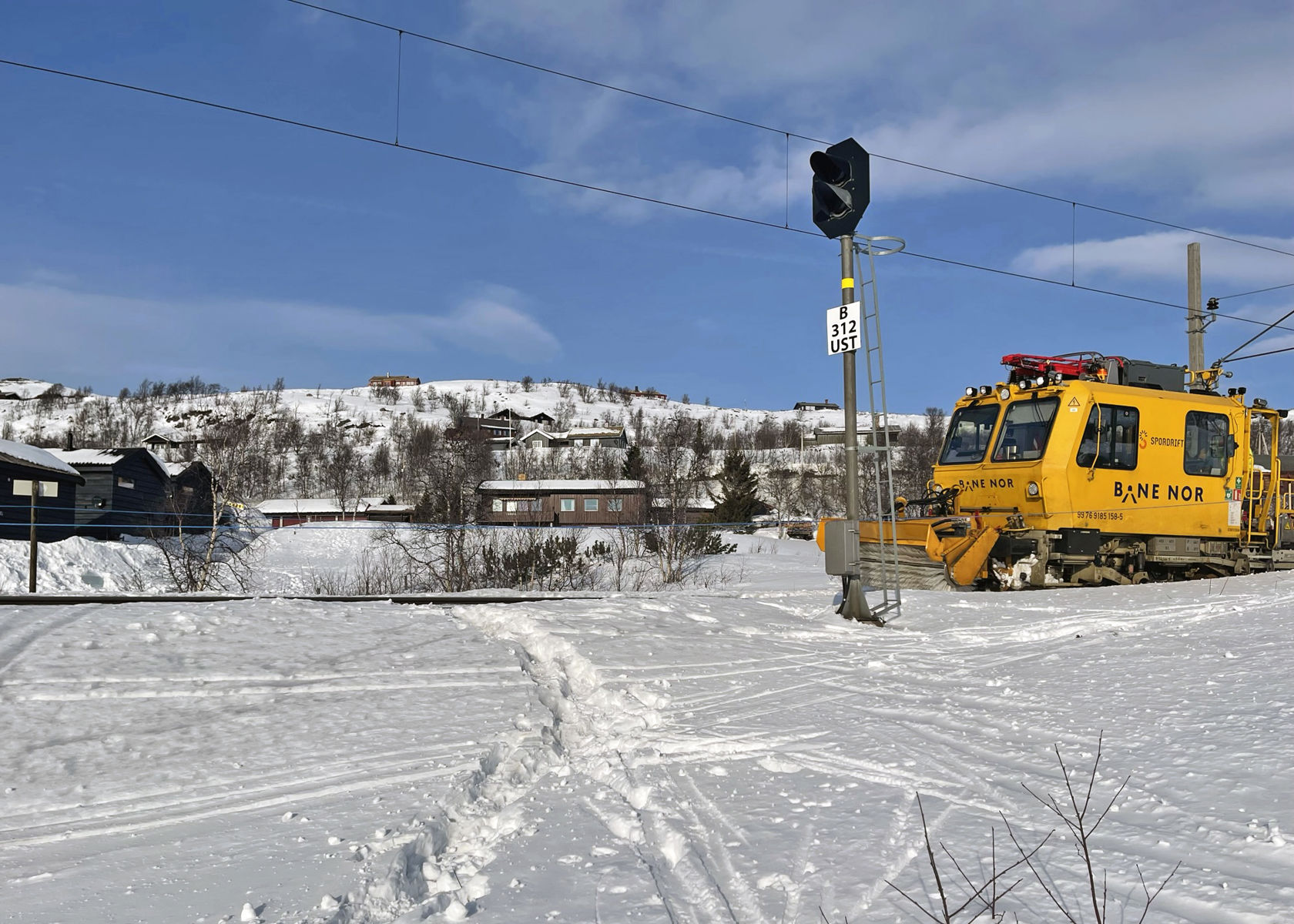 Spor i snøen krysser jernbaneskinnene. En gul arbeidsmaskin fra Bane NOR står på sporet.