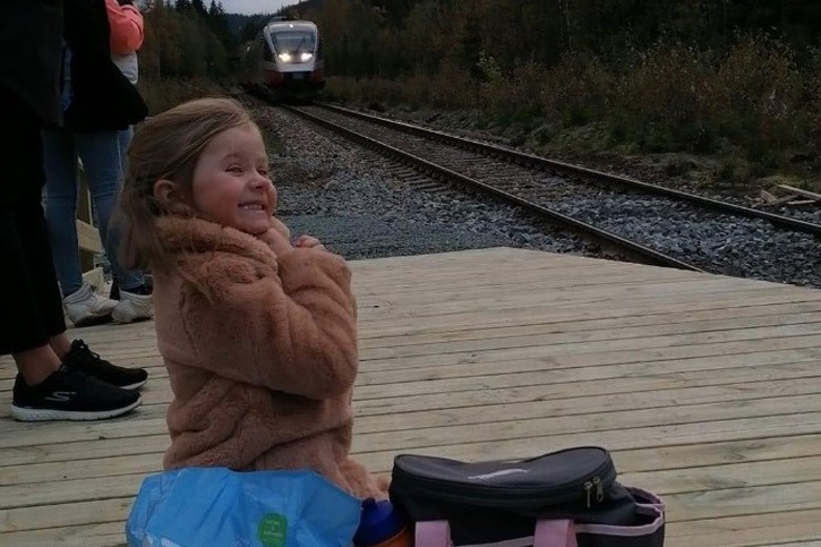 Ei lita jente sitter på en jernbaneplattform og venter på toget. Hun smiler og gleder seg. 