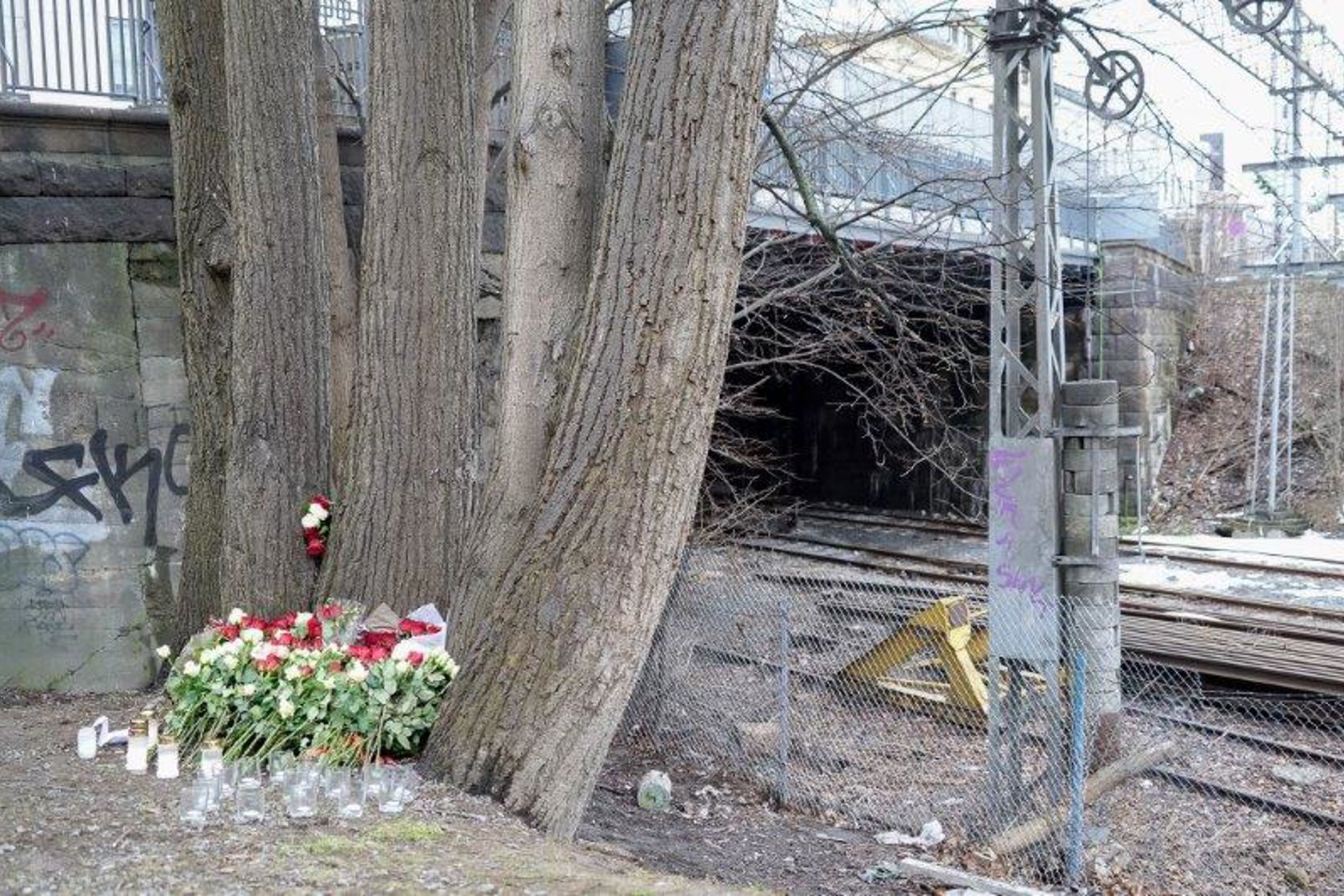 Et minnested med blomster og lys inntil et tre, foran et inngjerdet jernbaneområde