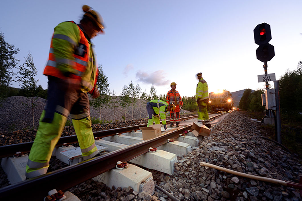 Folk i gule arbeidsklær jobber ute i skinnegangen. I bakgrunnen ser vi et arbeidstog.