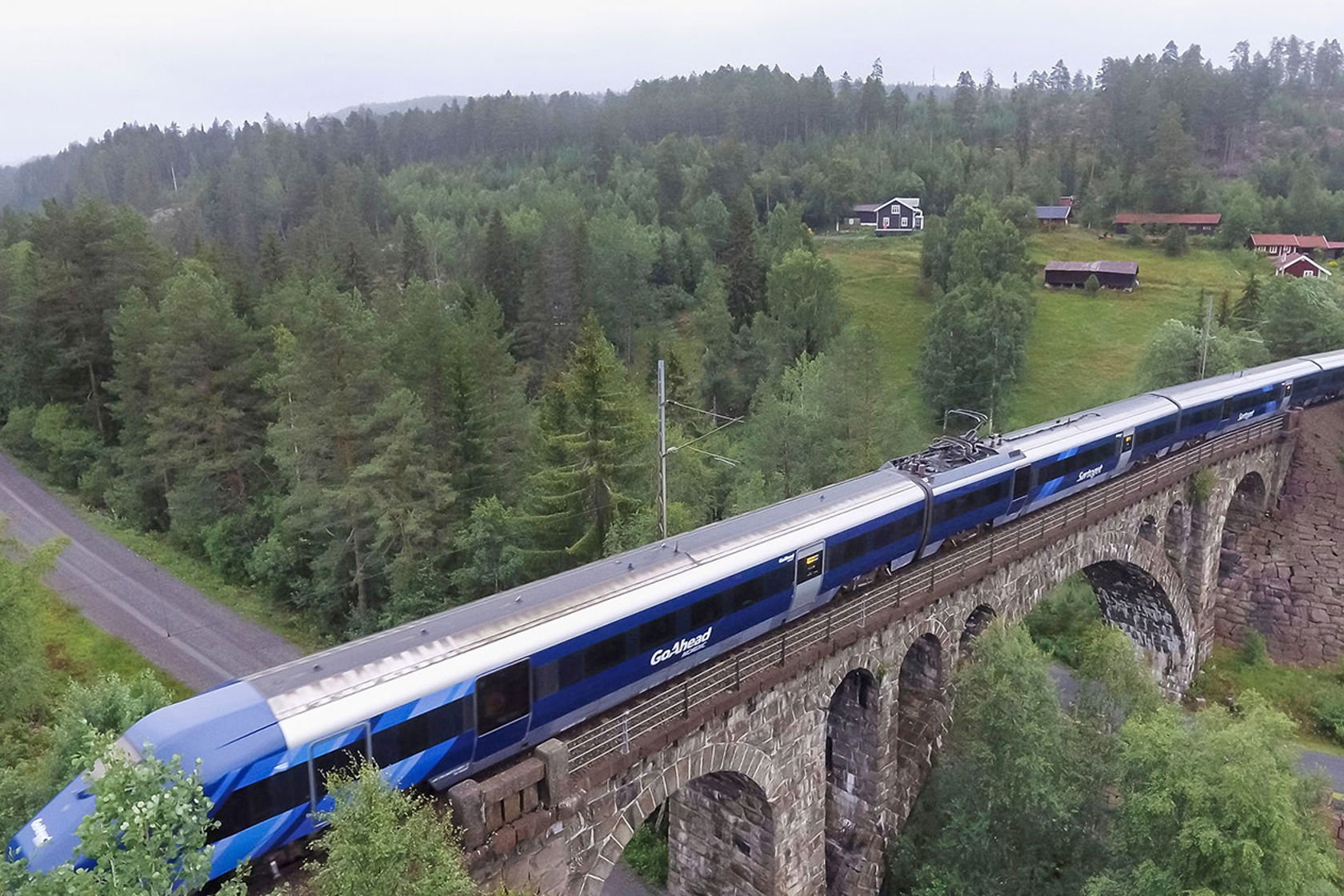 Dronefoto av blått tog som passerer over steinhvelvsbrua.