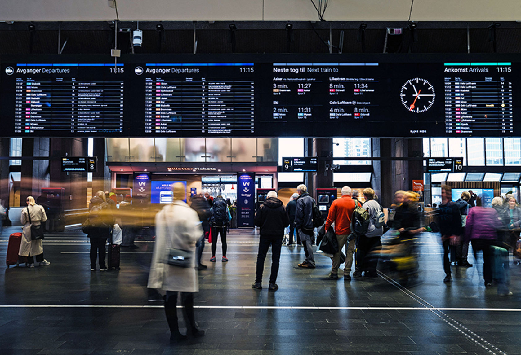 Folk som står under en infoskjerm på Oslo S som viser togtidene.