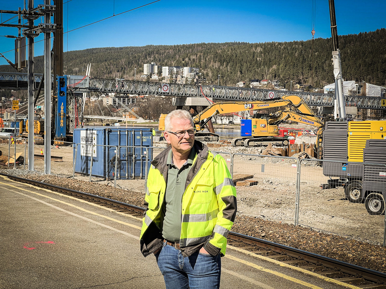 Mann i gul vernejakke står på plattform og ser mot høyre. I bakgrunn er anleggsområdet med maskiner.