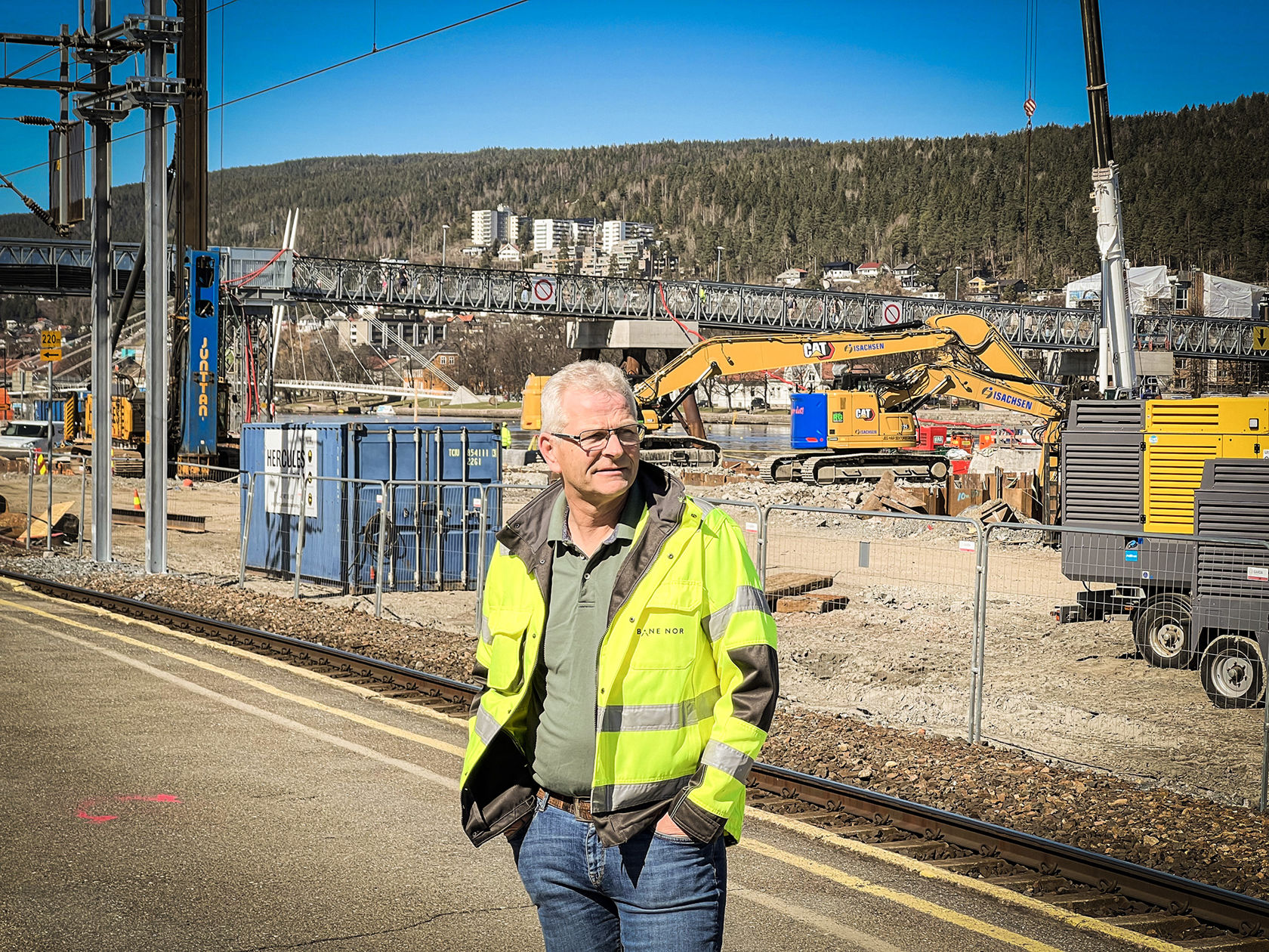 Mann i gul vernejakke står på plattform og ser mot høyre. I bakgrunn er anleggsområdet med maskiner.