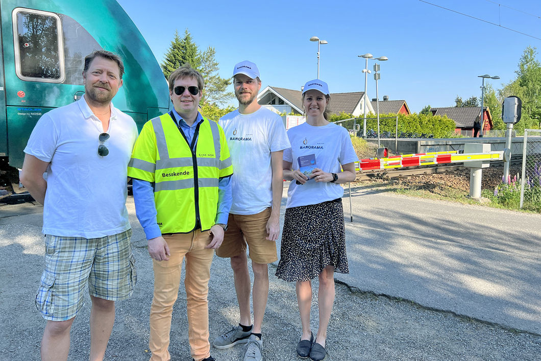 Personene står foran vegbommen, som er nede. Et Vy-tog passerer i bakgrunnen.