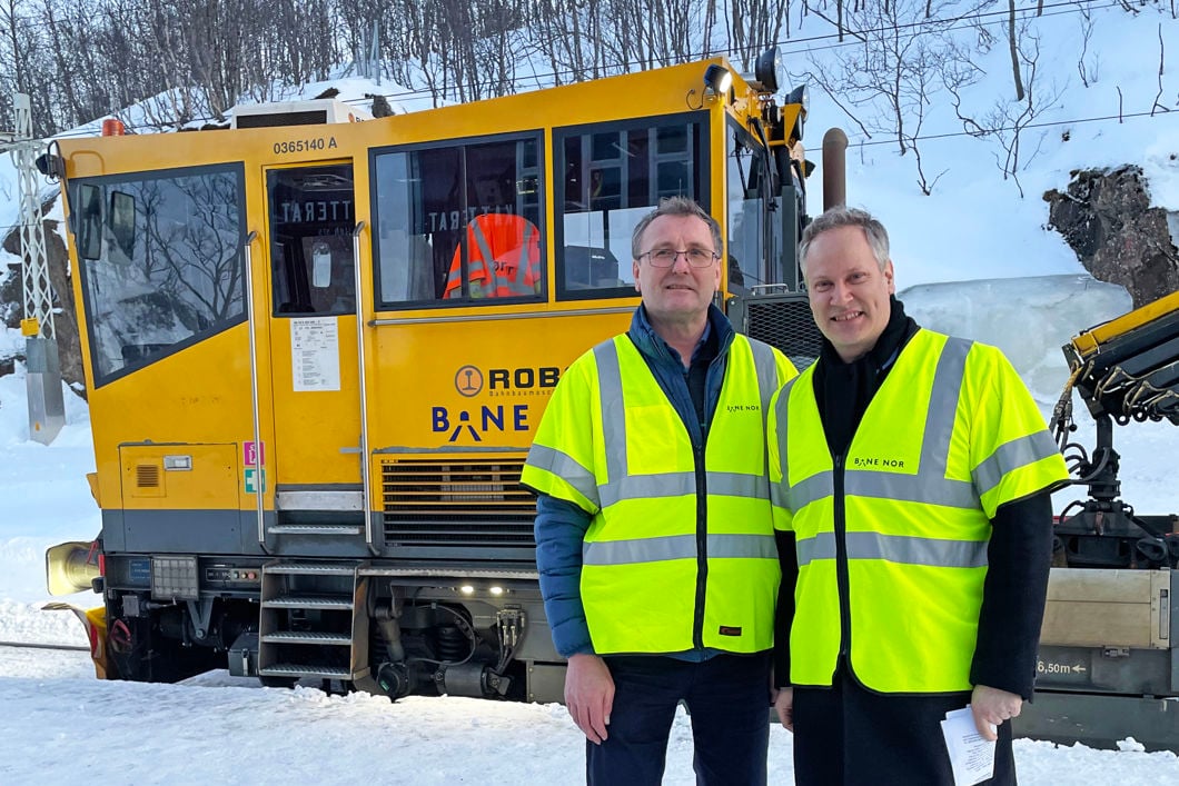 To menn i gule vernejakker står foran en gul arbeidsmaskin. Det er snø og vinter.