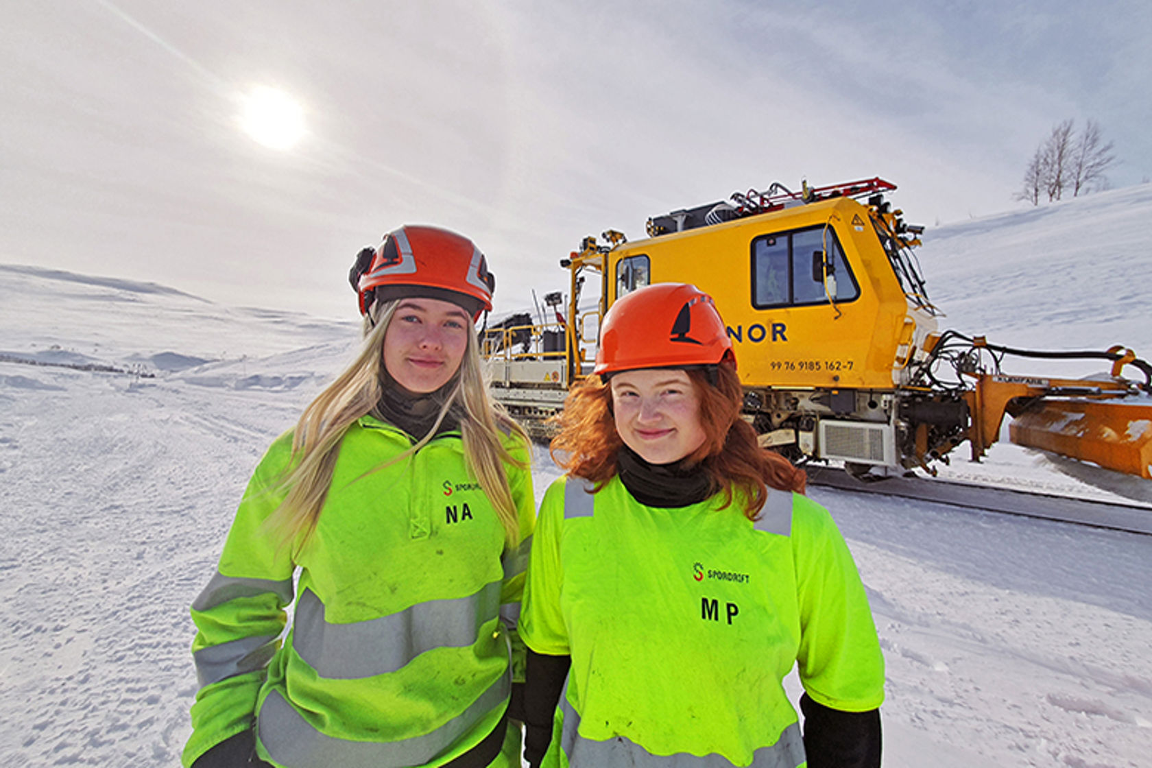 Bilde av to unge kvinner som står i arbeidstøy og hjelmer foran et gult arbeidstog på Saltfjellet.