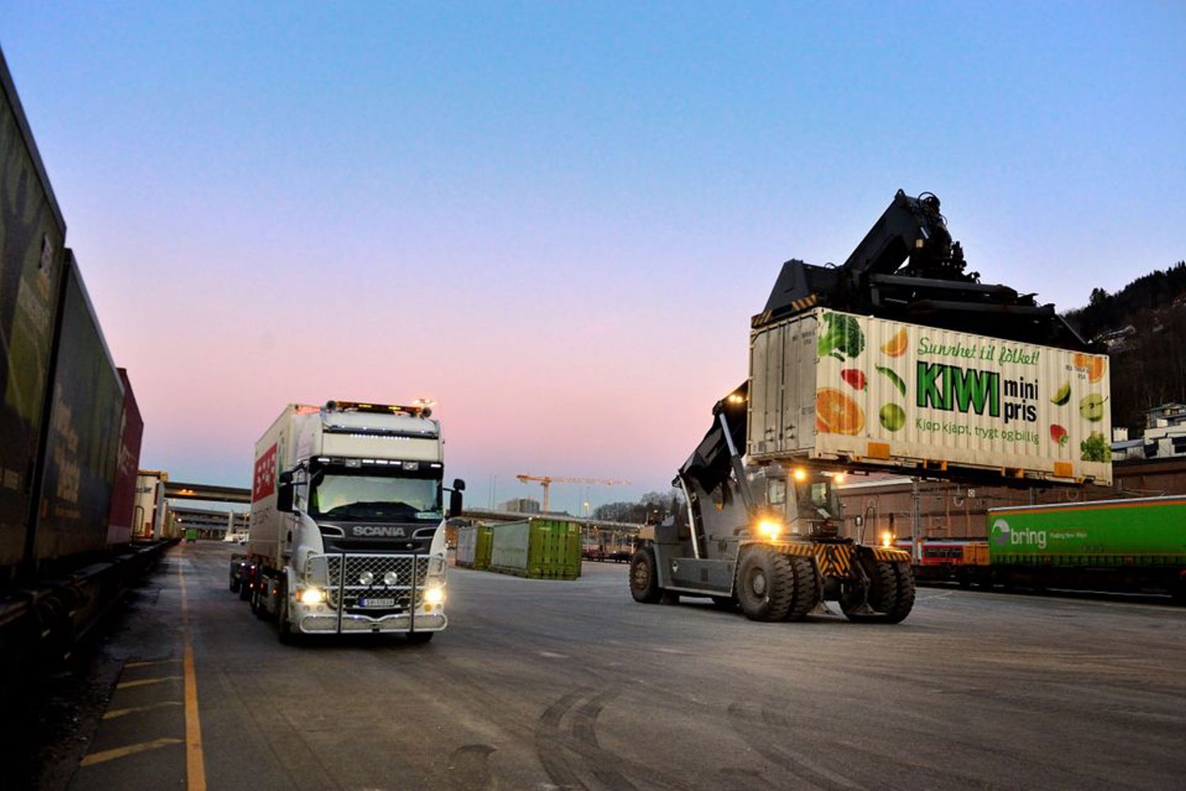 Bildet er tatt på en godsterminal hvor en stor truck laster av en trailer og på et godstog. 
