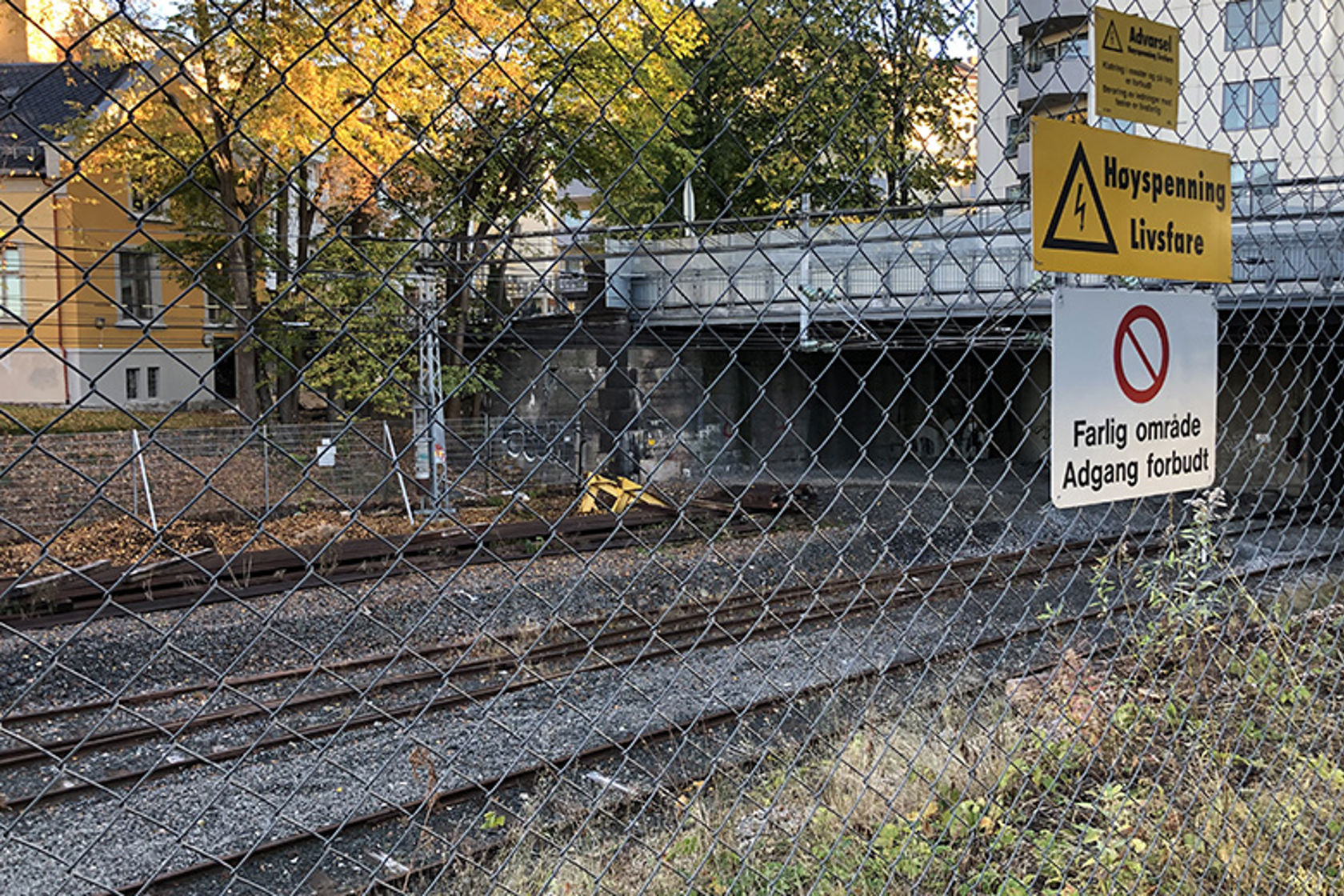 Bilde av et gjerde med flere sikkerhetsadvarsler foran et område med jernbanespor.