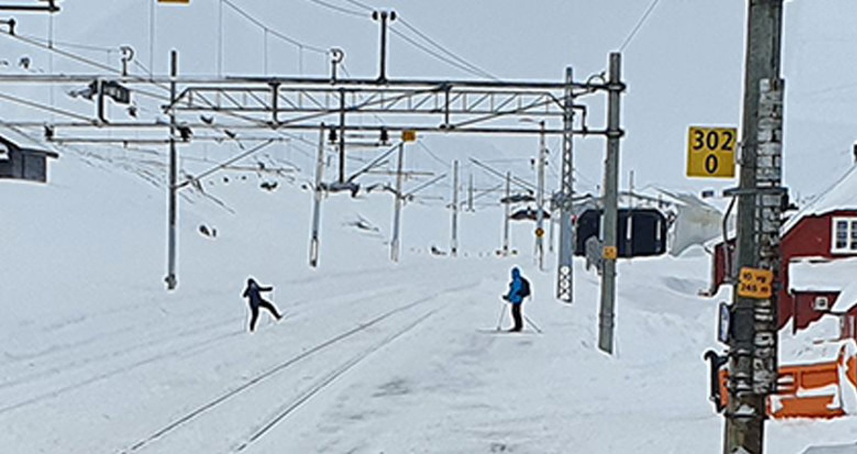 To skigåere tråkker over jernbanens spor inne på en snødekt Finse stasjon.