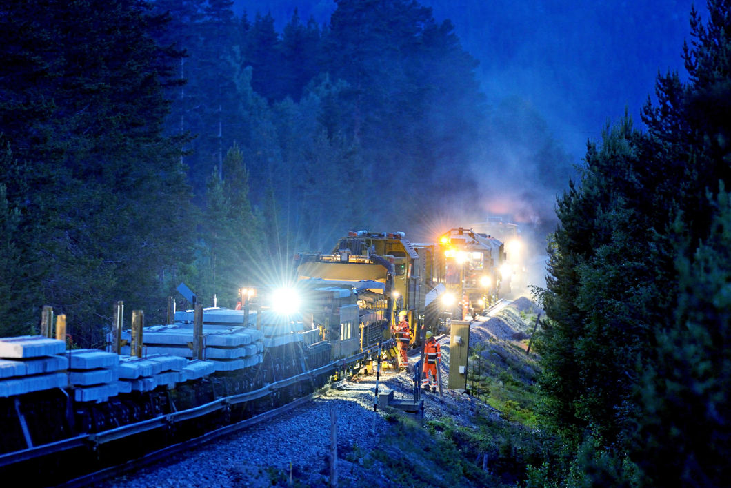 Et stort arbeidstog lyser opp en mørklagt skog under arbeid på skinnegangen.