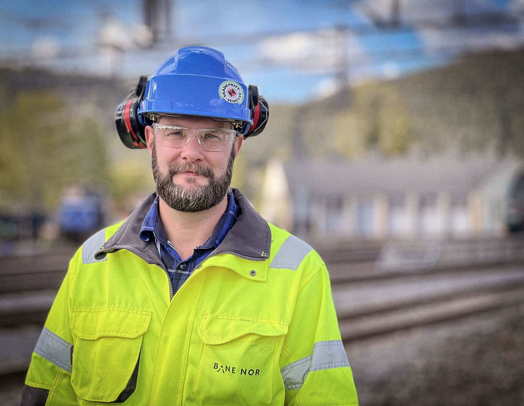 Mann med gult vernetøy og blå hjelm. Jernbaneområde i bakgrunnen.