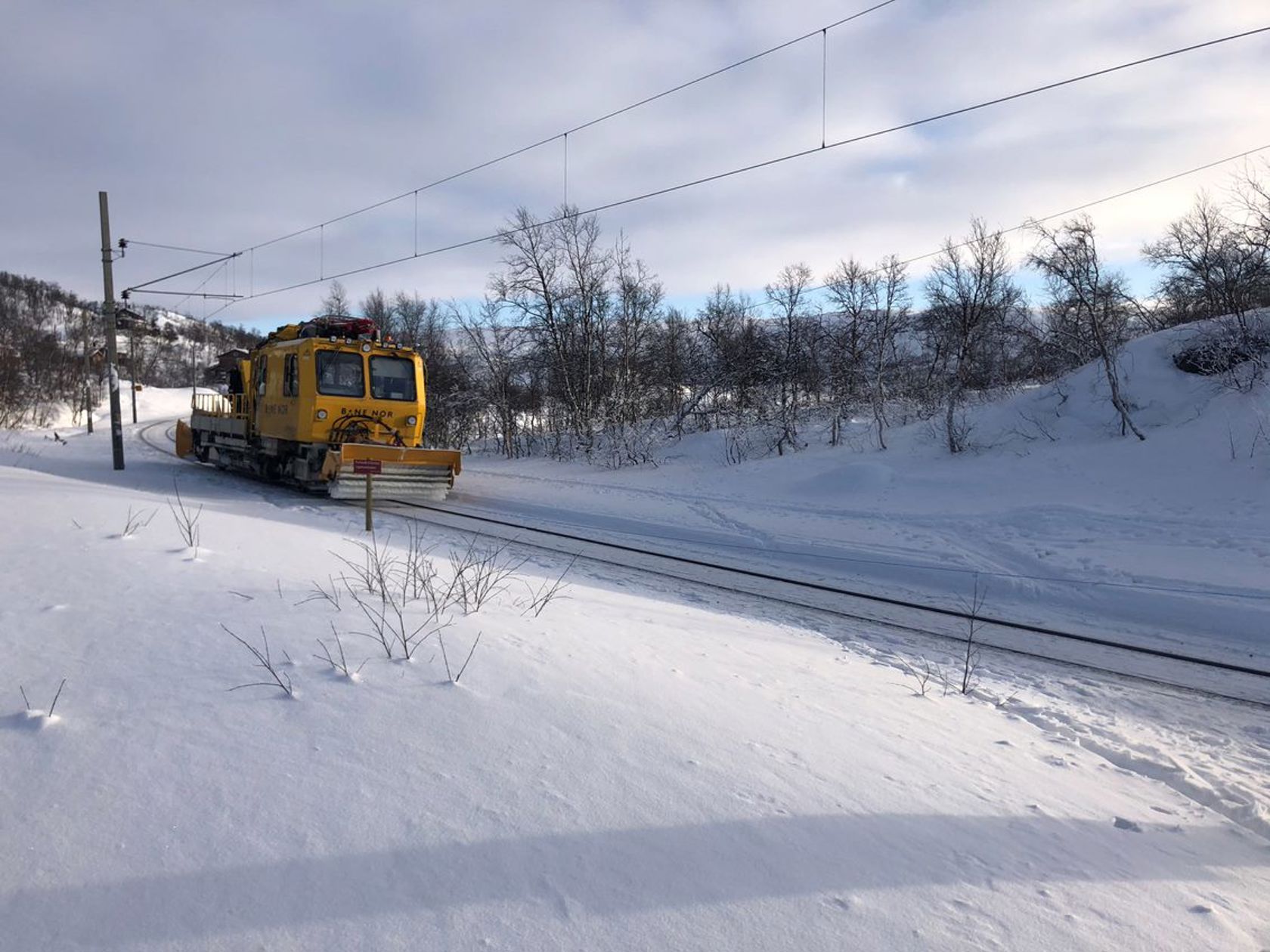 Bilde av et gult arbeidstog som kjører gjennom et snødekt fjellandskap. 