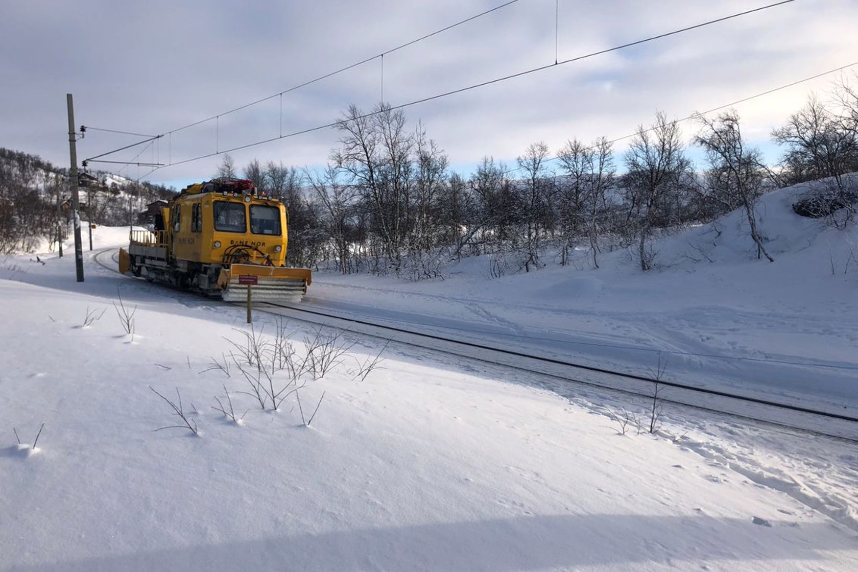 Bilde av et gult arbeidstog som kjører gjennom et snødekt fjellandskap. 