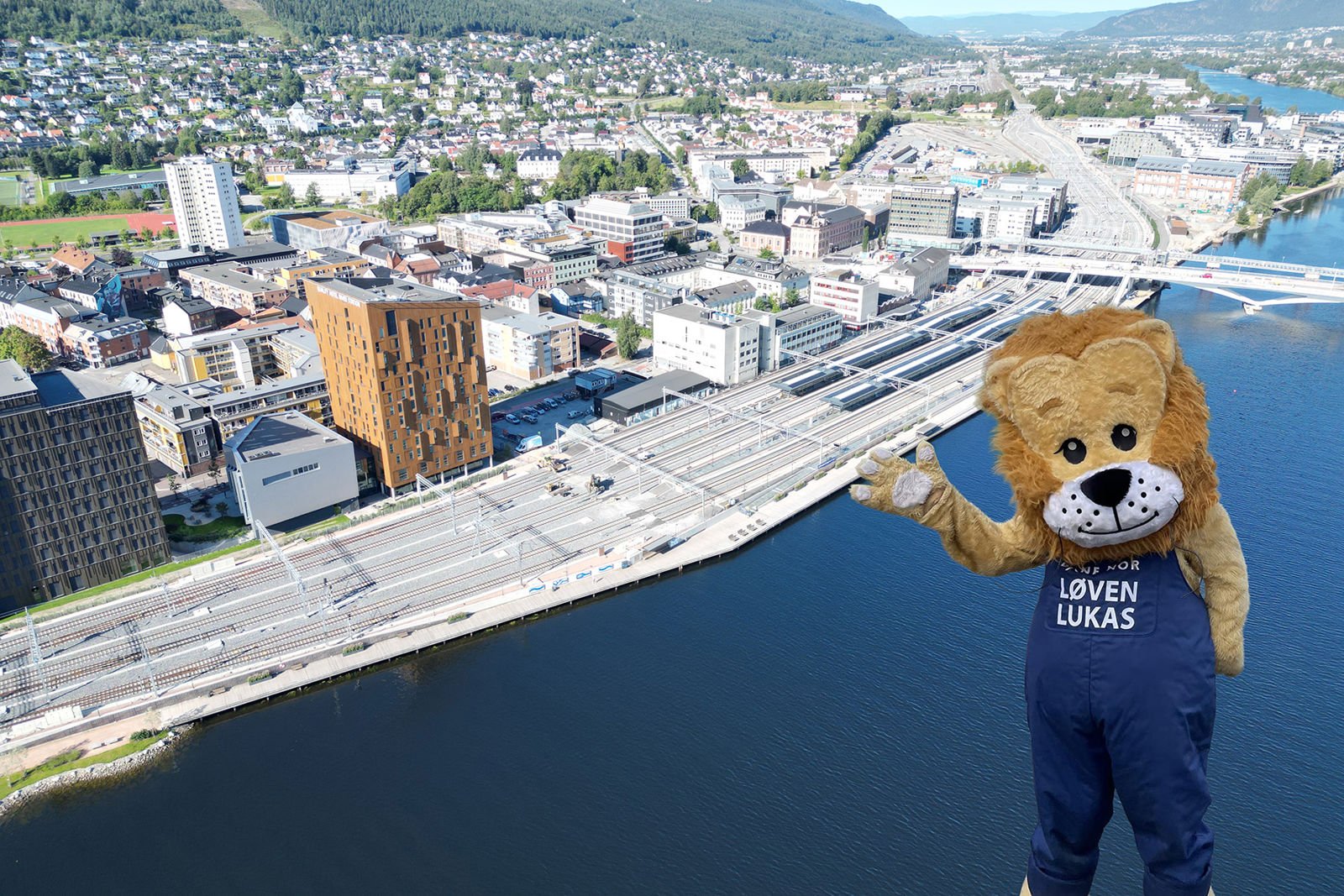 Montasje: Maskoten Løven Lukas vinker foran dronefotografi av Drammen stasjon,
