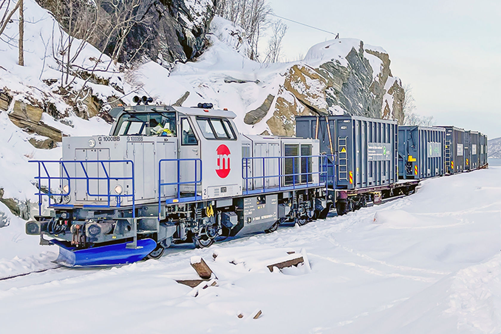 Et godslokomotiv med godsvogner bak står på et togspor. Det er vinter. 
