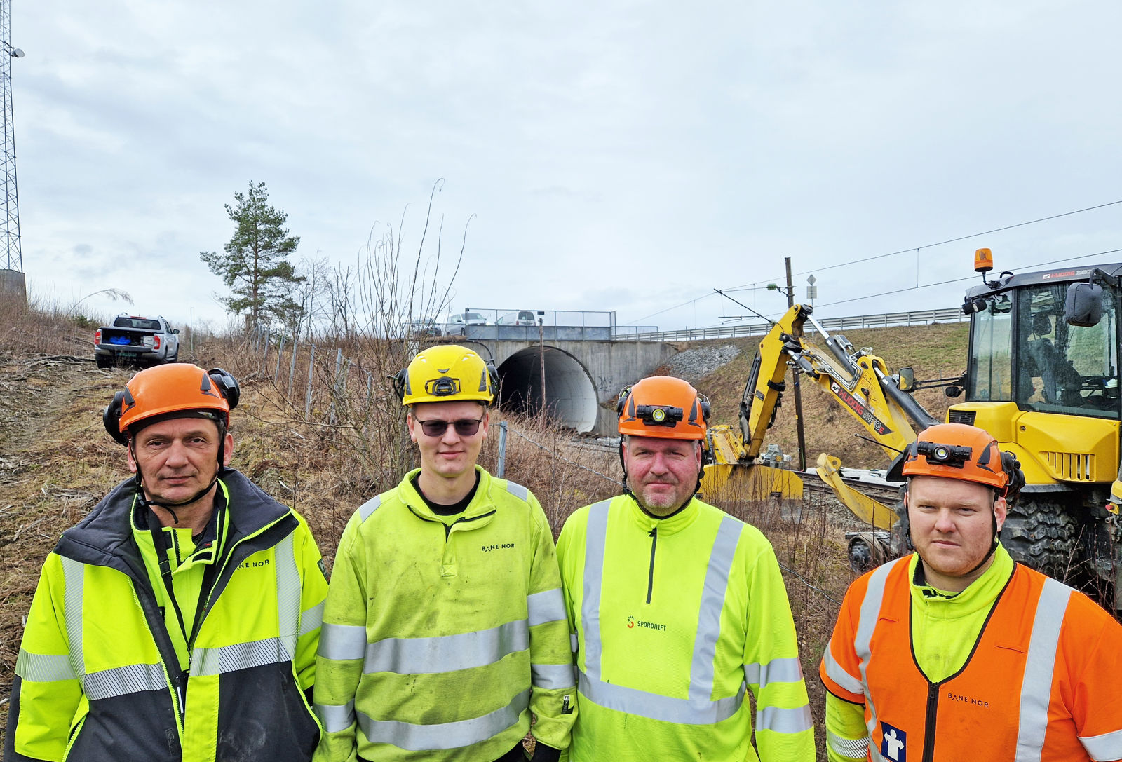 Fire menn i verneklær med en arbeidsmaskin i bakgrunn.