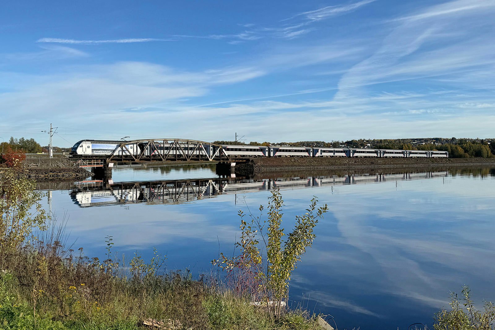Høstbilde av tog på dagens enkeltspor med bru og fylling, toget speiler seg i vannet.