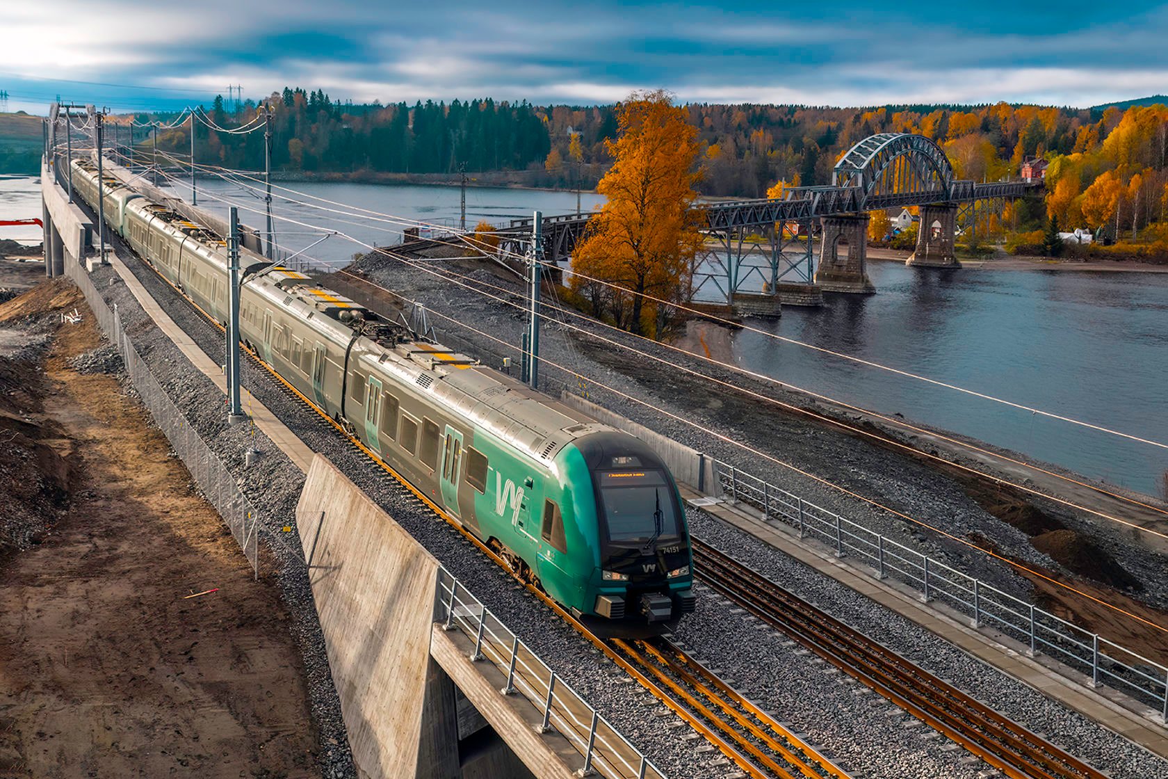 Et grønt Vy-tog med Vorma, gammel og ny jernbanebru i bakgrunnen.