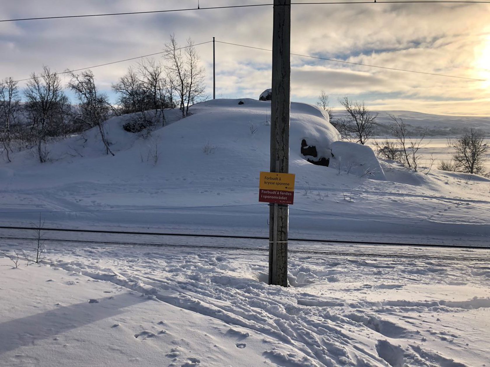 I et snødekt terreng står det en stolpe med advarsel om å krysse jernbanen. Ved siden av går skispor som krysser jernbanen.