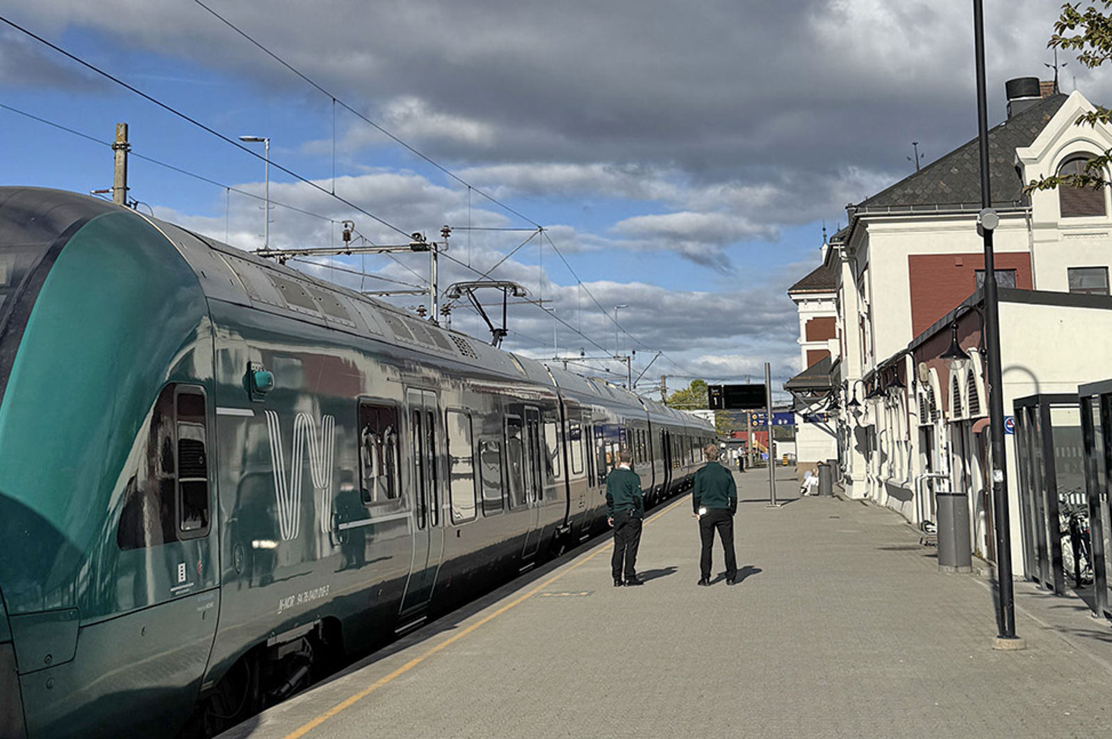 Grønt Vy-tog til venstre, personer på plattformen, stasjonsbygningen og sykkelparkering til høyre.