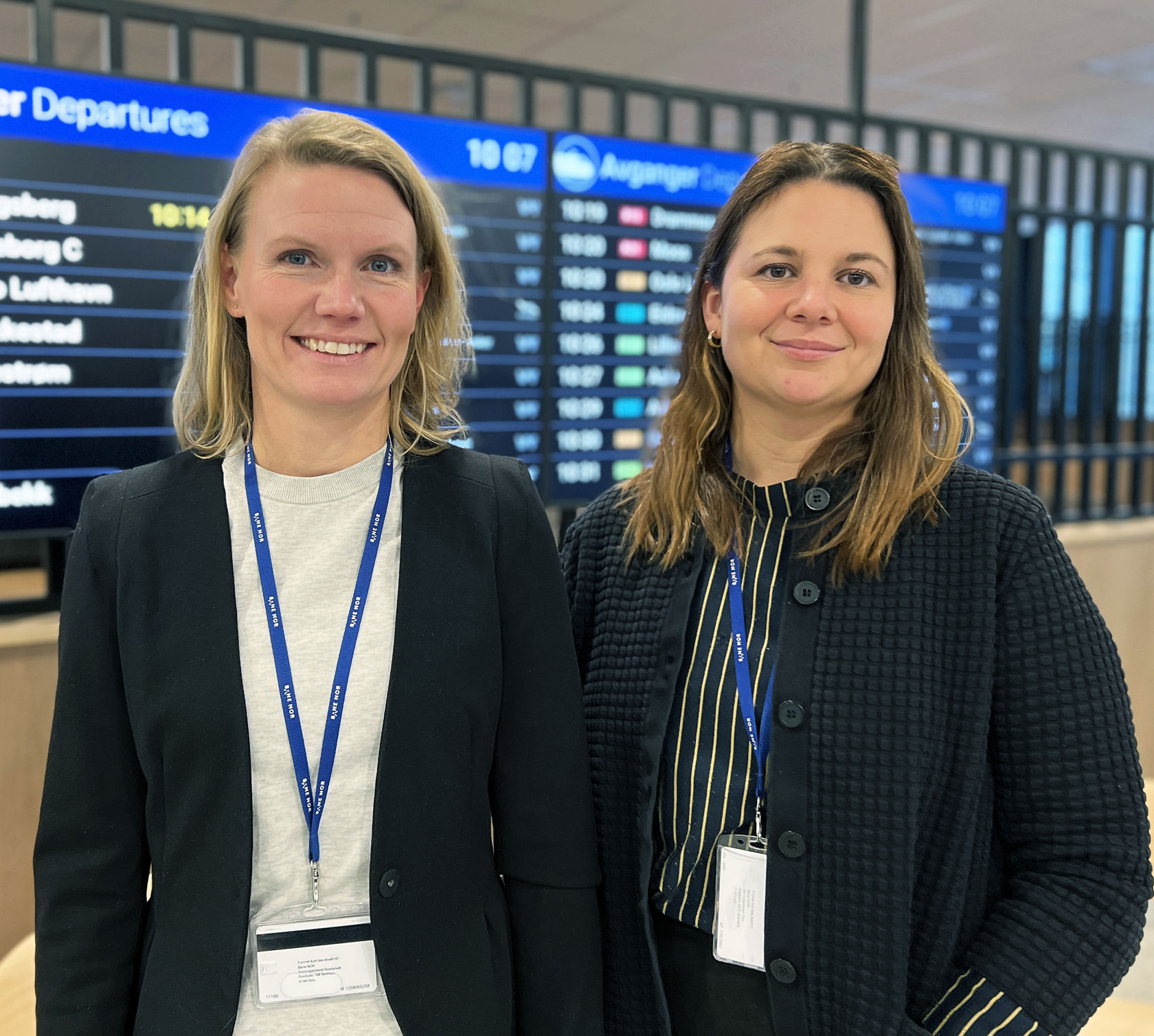 Bildet viser to kvinner som smiler og står foran en digital informasjonstavle.