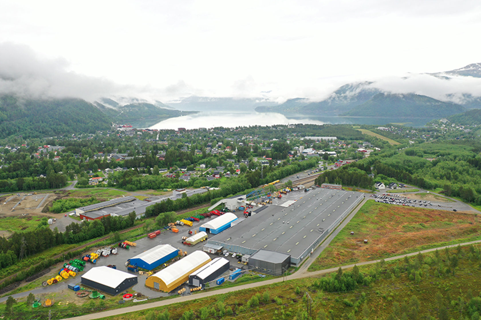 Bilde tatt ovenfra av stor bygning i et industriområde, med fjell og fjord i bakgrunn.