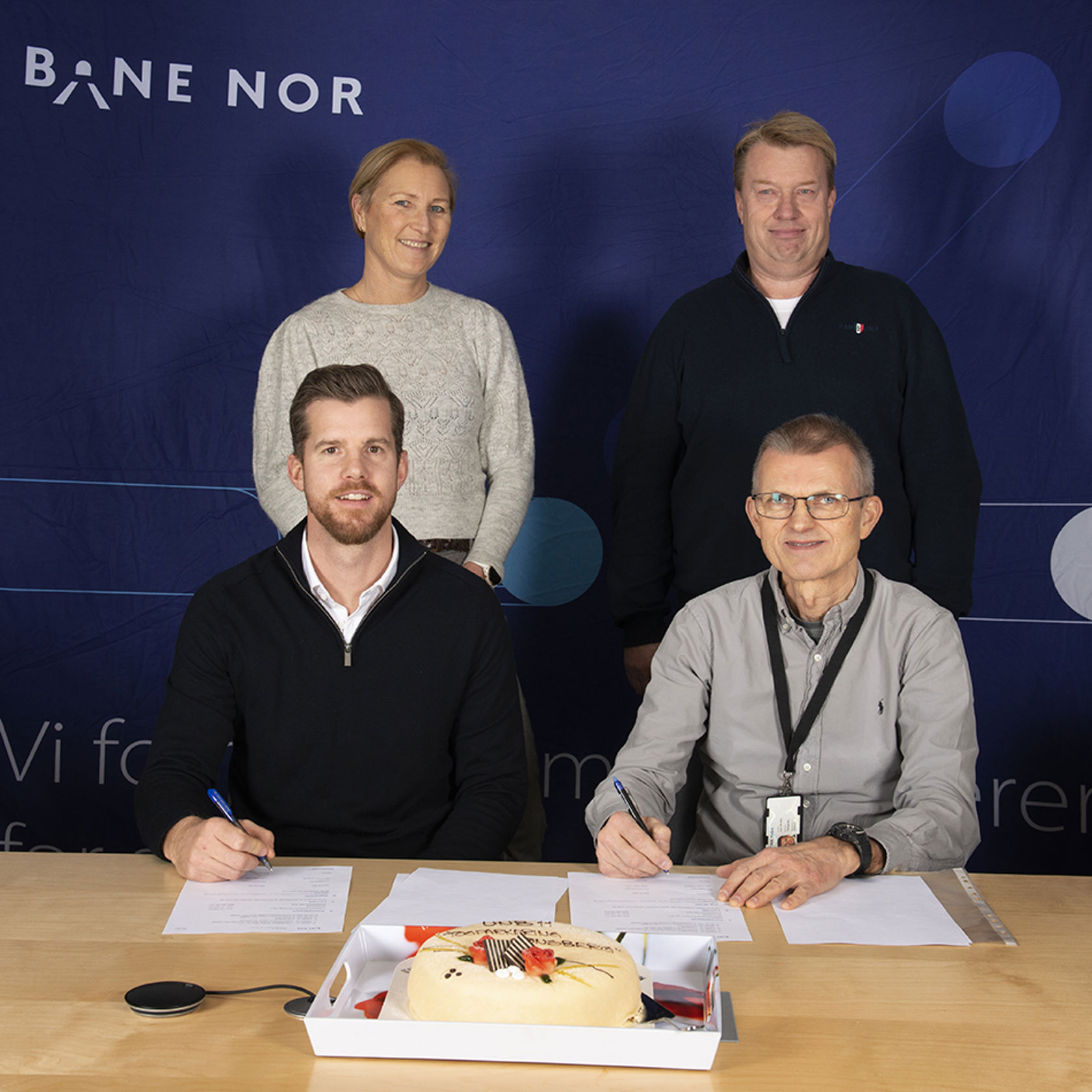 To menn signerer kontrakt, en kvinne og mann står bak. Foran på bordet står det en bløtkake
