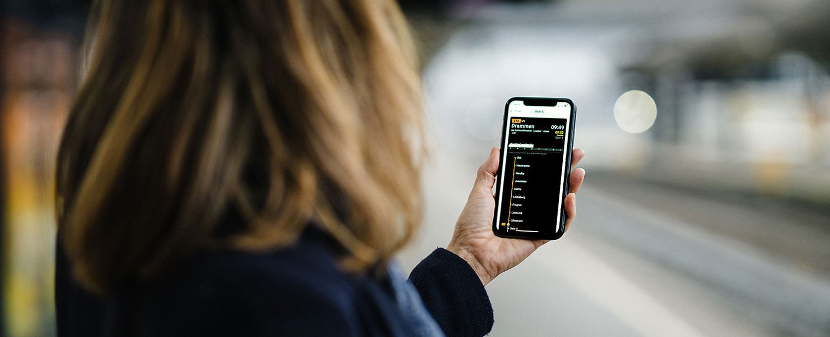Reisende på en plattform holder mobiltelefon med NÅ-appen til Bane NOR.