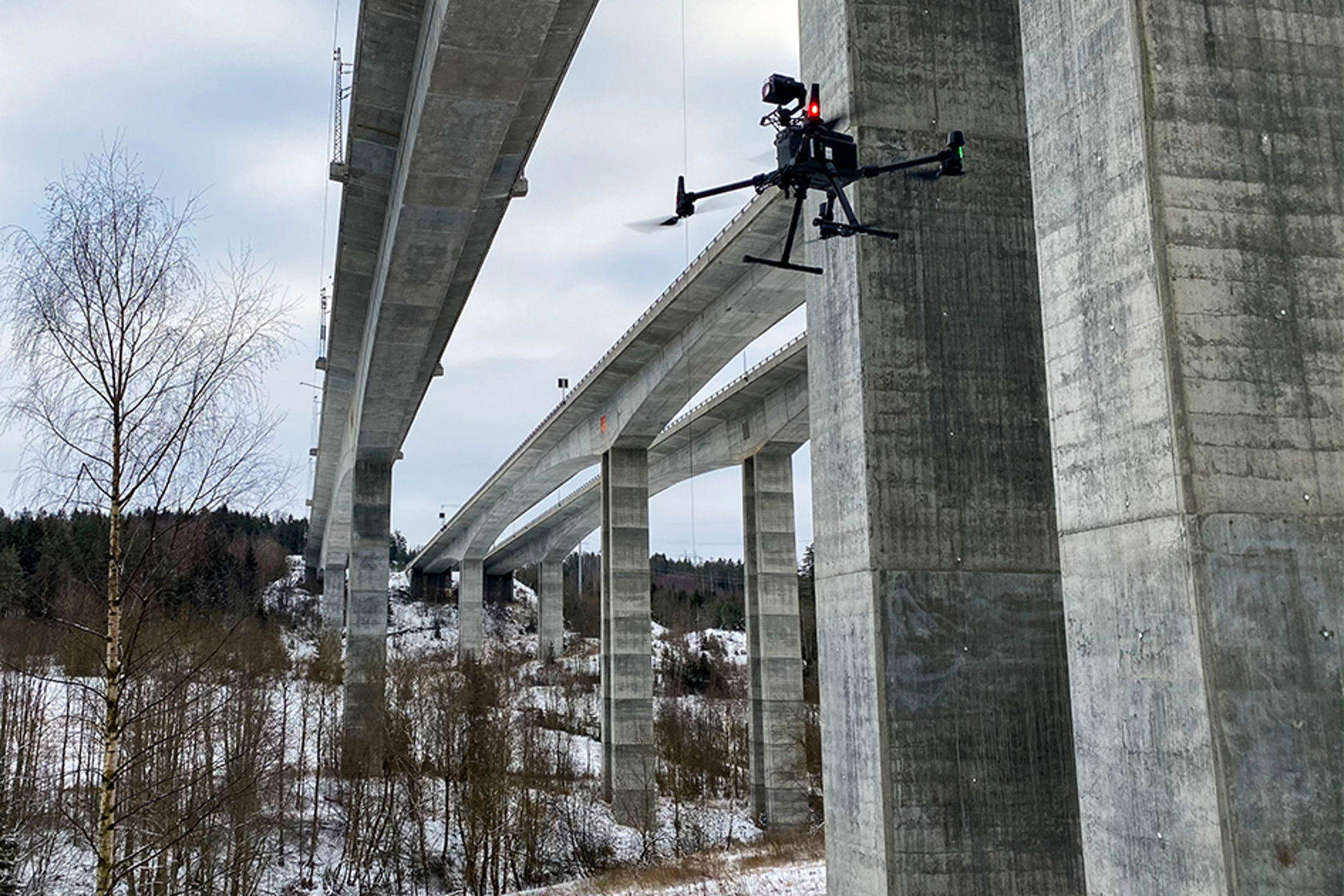 Bildet viser en drone som flyr rundt betongbeina på en bru.