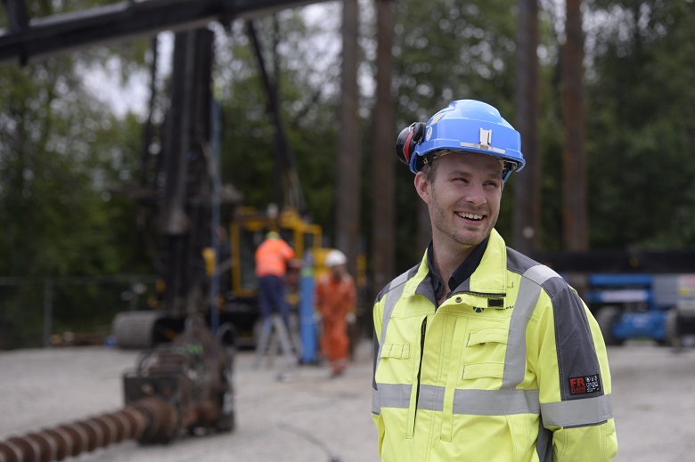 Christer Larsen, kontraktsrådgiver, ute i felt. Christer er kledd i verneutstyr, gul jakke og blå hjelm, og står foran byggeområdet han jobber med. 