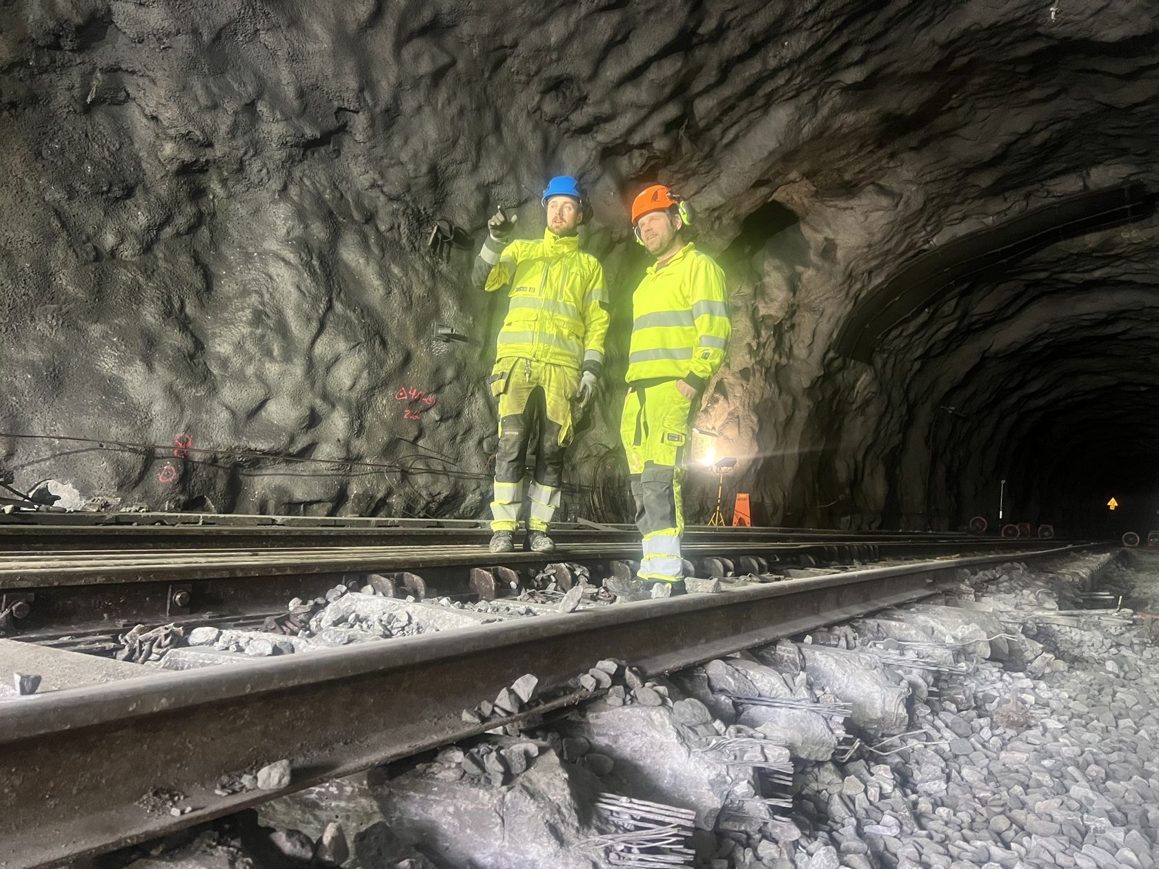 To arbeidsmenn i gule klær står på jernbaneskinner i en tunnel. En peker innover i tunnelen.