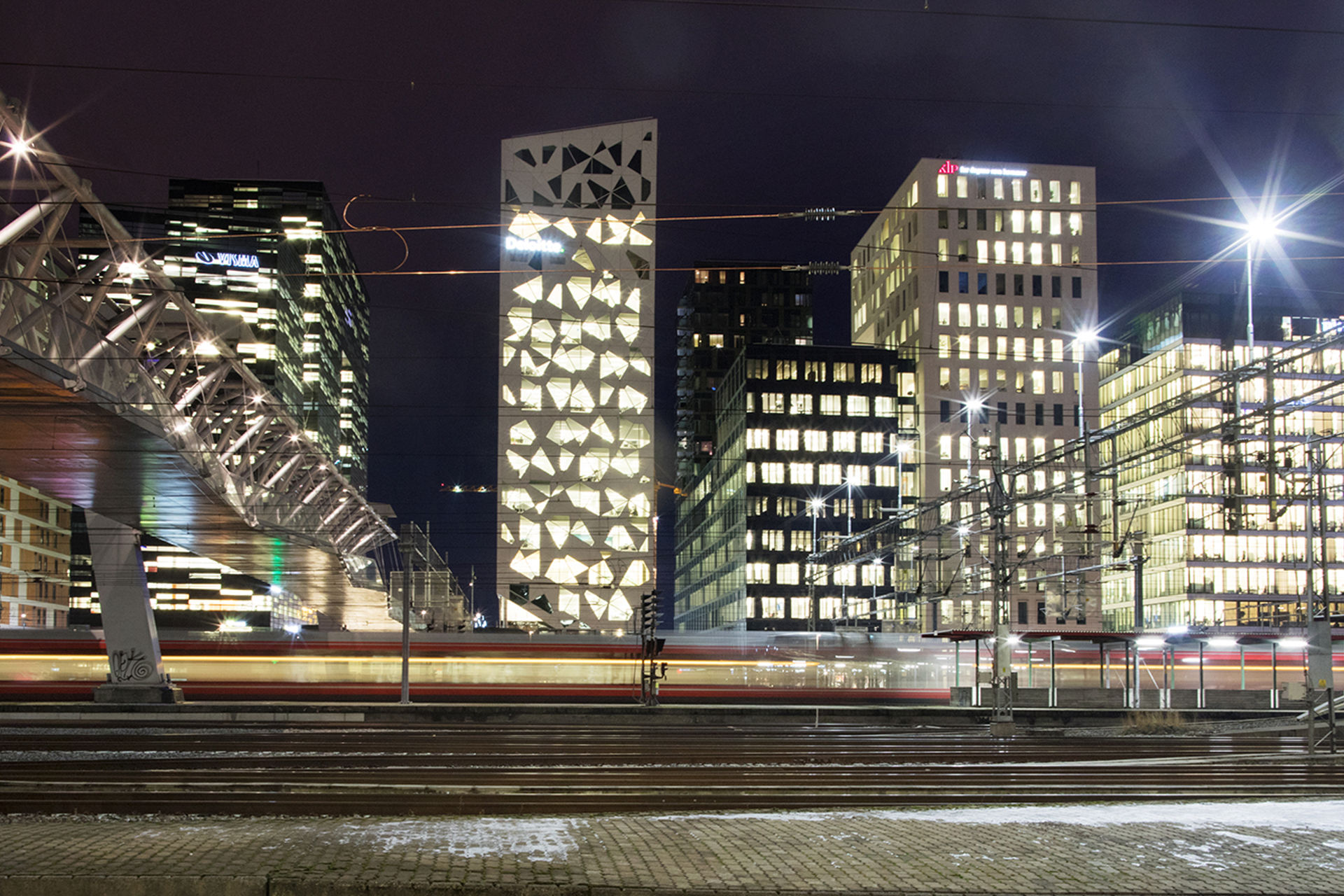 Høye lysende bygninger i mørket, med togskinner forran