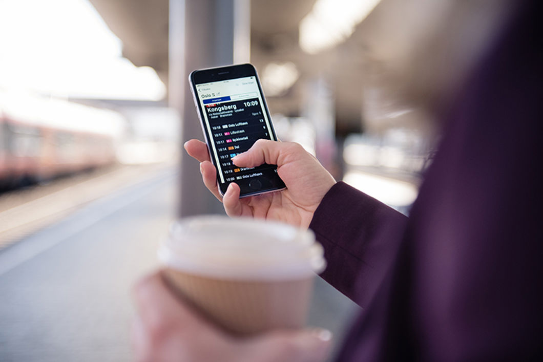 En person står på perrongen og sjekker avganger i NÅ-appen på mobilen.